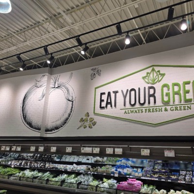 White faux brick wall panels serving as a textured backdrop for "Eat Your Greens" signage in a supermarket produce section.