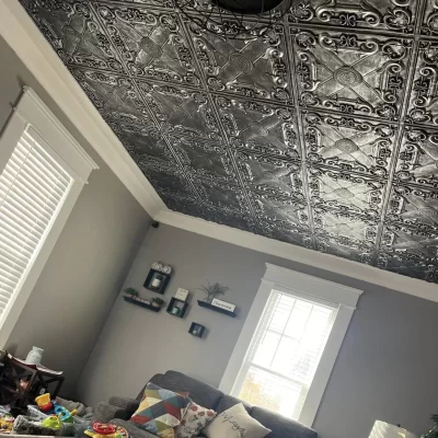Angled view of TD22 Rustic Faux Tin ceiling tiles in Aged Silver meeting white crown molding above grey living room walls